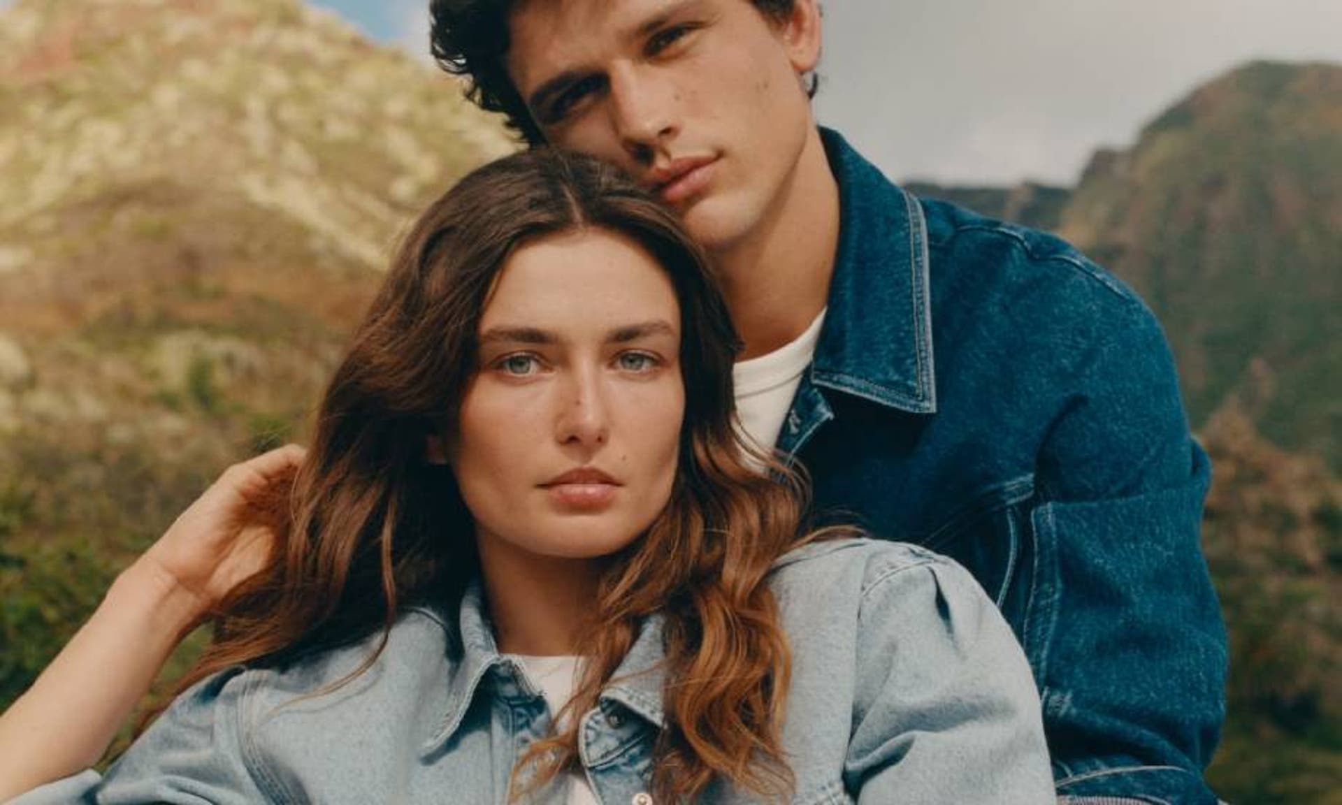  A brunette couple wearing matching denim jackets and white tops from Mango, sat close together in front of a rural backdrop. 