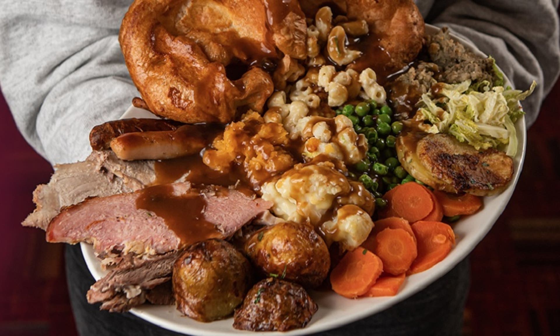  A picture of a Toby Carvery roast featuring beef, gammon, roast potatoes, mash potatoes, yorkshire puddings, sausages, mac and cheese and vegetables served on a plate with gravy held by someone wearing a grey jumper. 