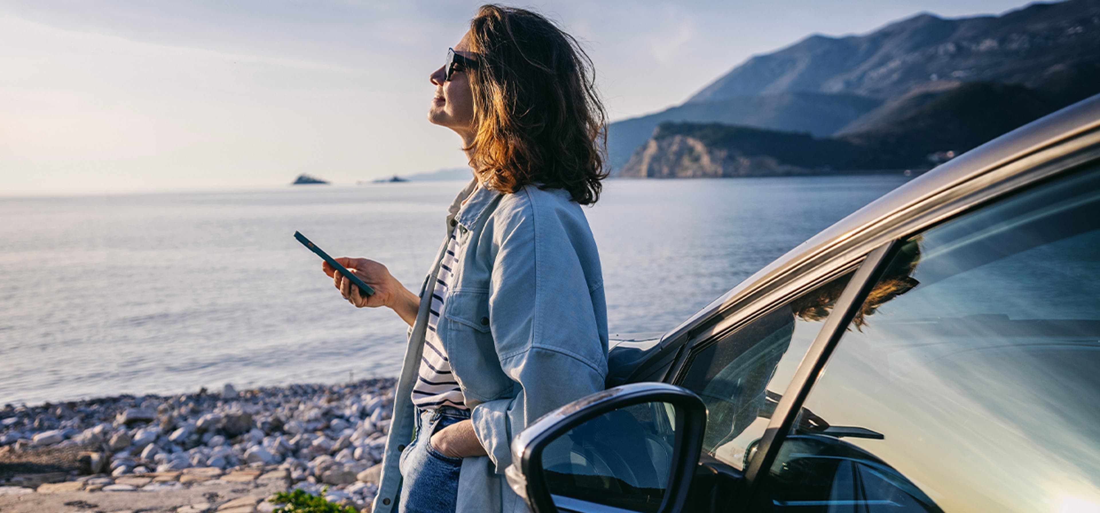 Woman leant against her car, parked up on a beach front with the sun shining
