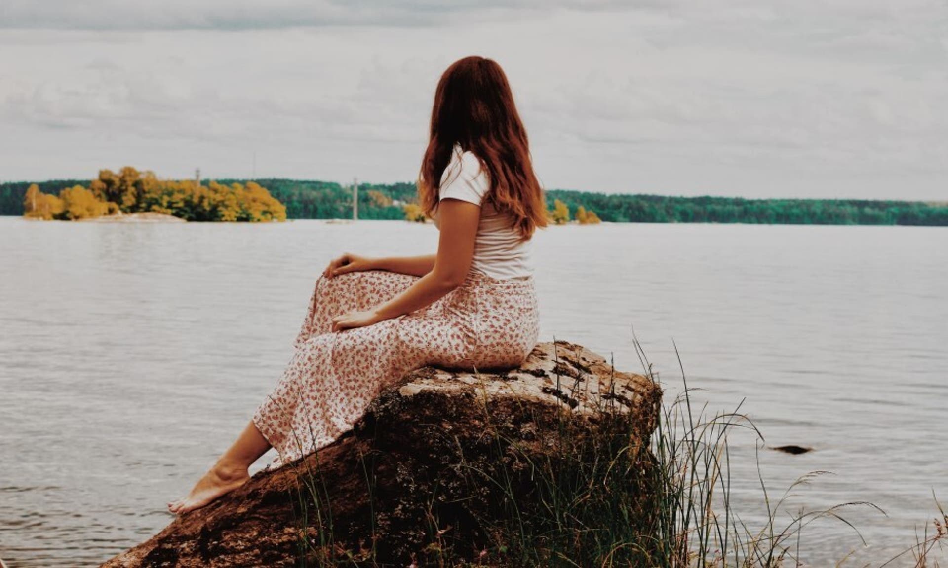  image of a woman sat on a rock, wearing a white top with a red and white floral skirt and looking out over a scenic lake. 