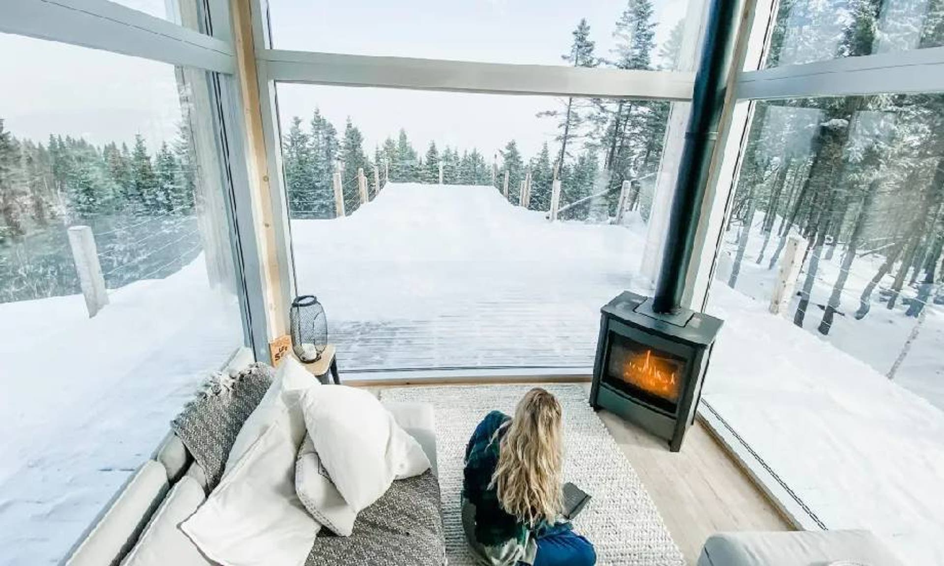  A woman sat in a glass property with a log burner, surrounded by a snowy backdrop with snow covered trees. 