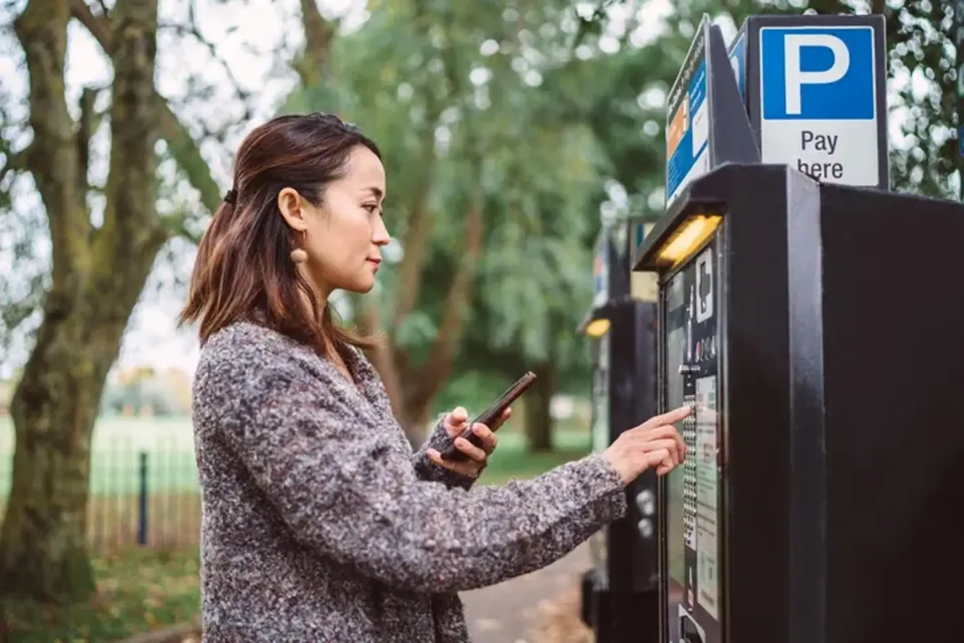  woman making contactless payment with smartphone at parking payment machine in city street 