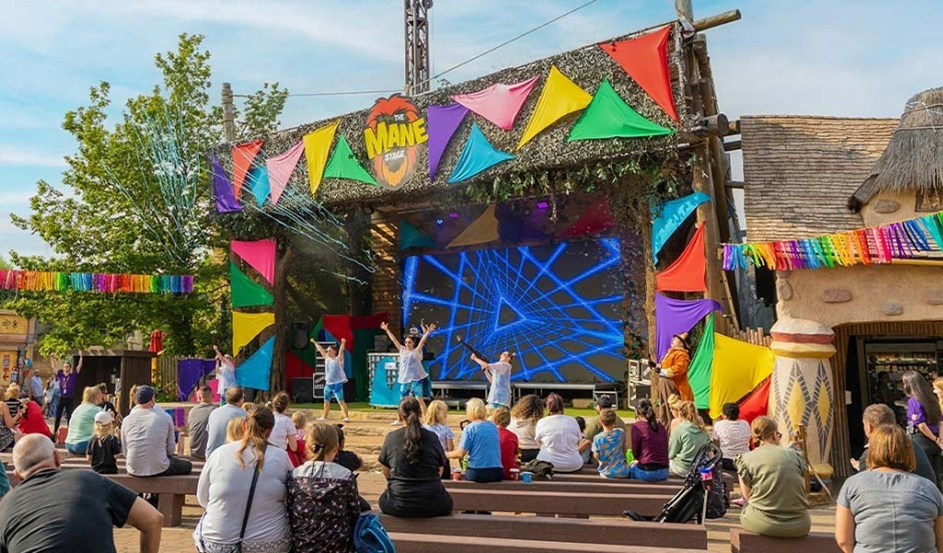  An image of people sat on benches watching performers perform on a stage at Chessington World of Adventures on a bright sunny day. 