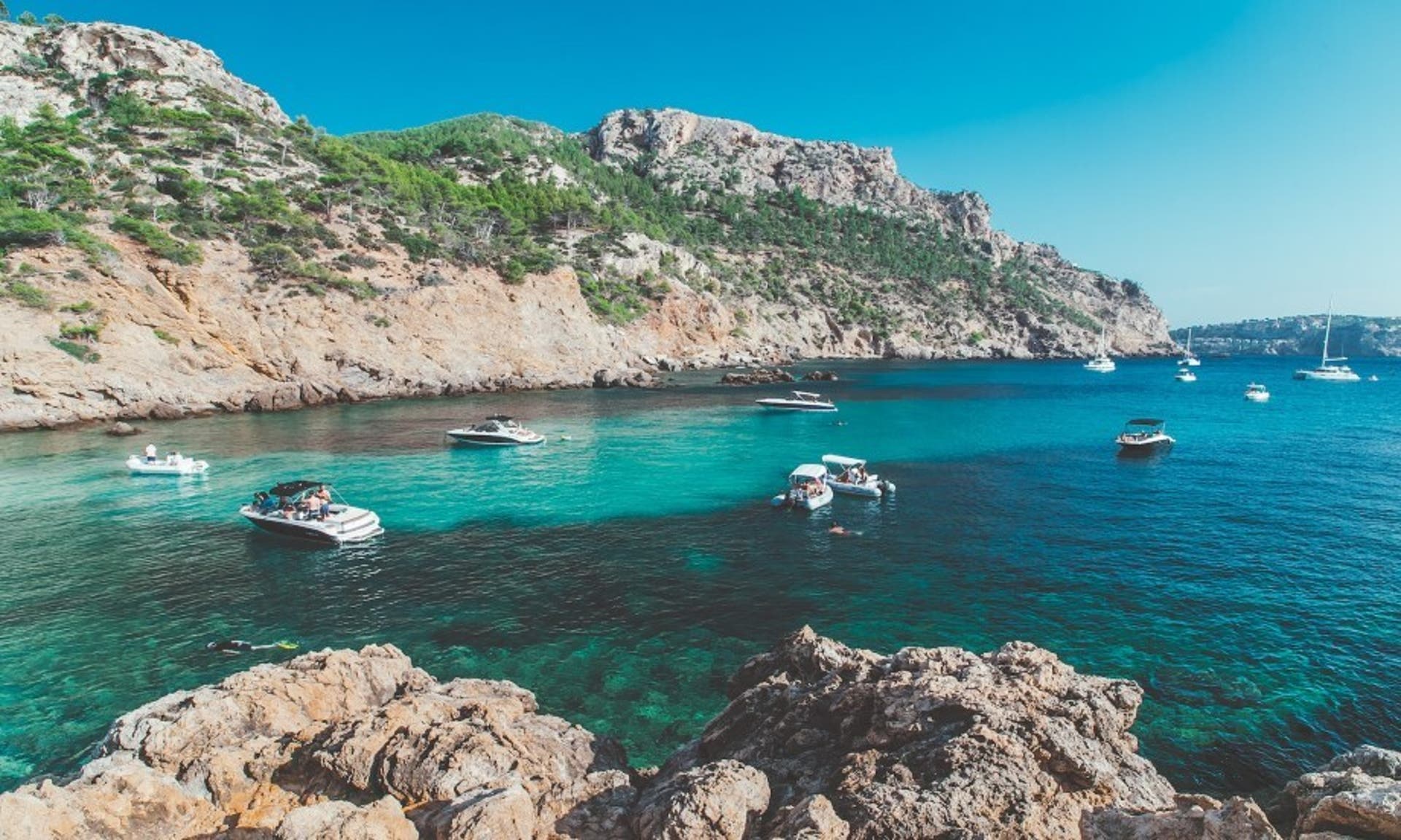  image of a coastal alcove in Mallorca, with idyllic light blue sea and people jumping off moored boats into the water 