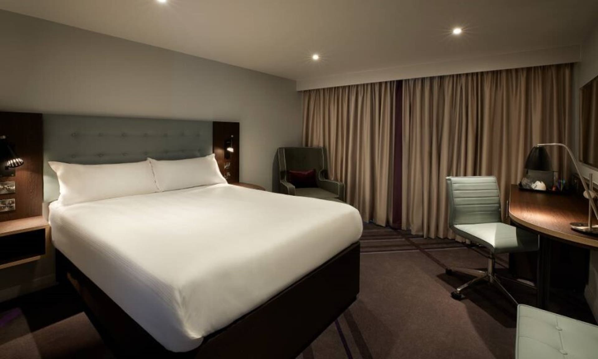 A picture of a Premier Inn room showing the bed, armchair and desk with a chair and desk lamp.