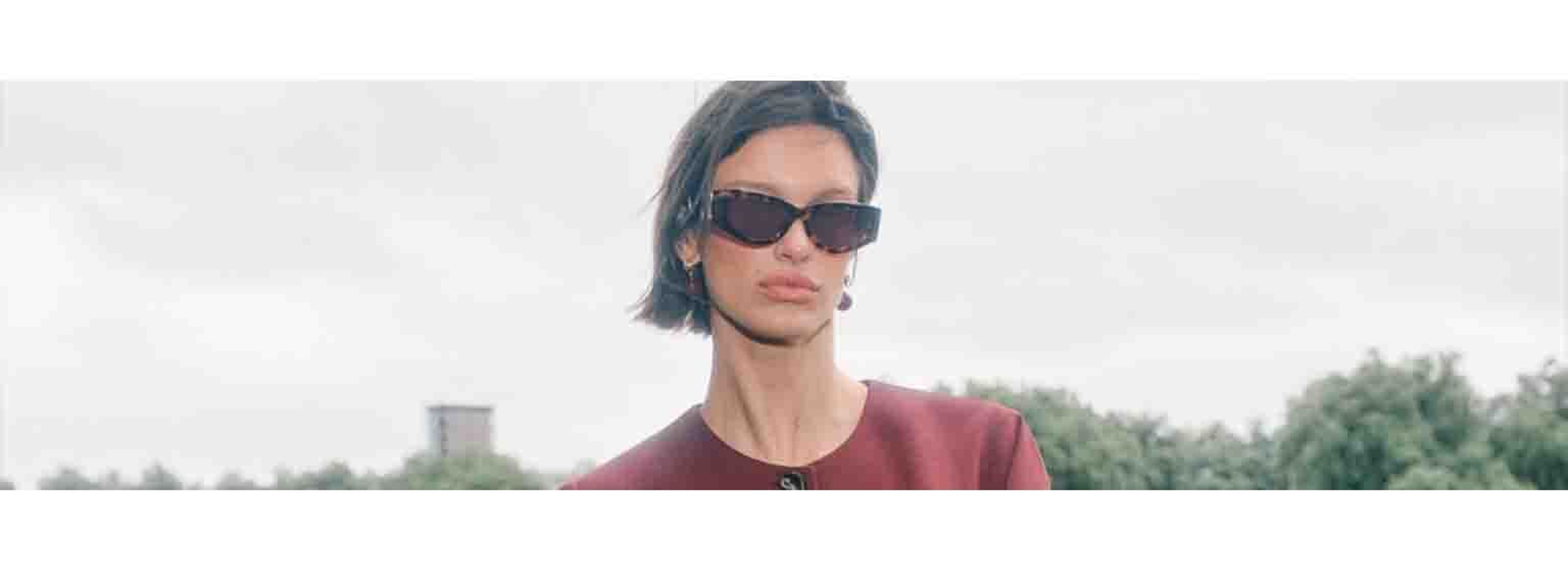 women in centre of frame from the shoulders up wearing sunglasses and red blazer, sky behind her
